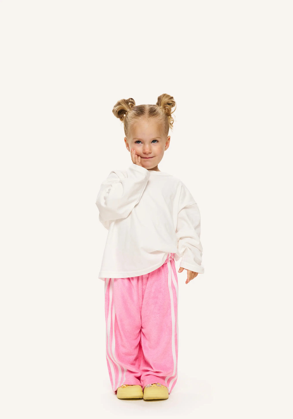 A young child with two buns of hair stands against a plain off-white background, wearing a white long-sleeve top, pink velour pants with white side stripes, and yellow shoes.