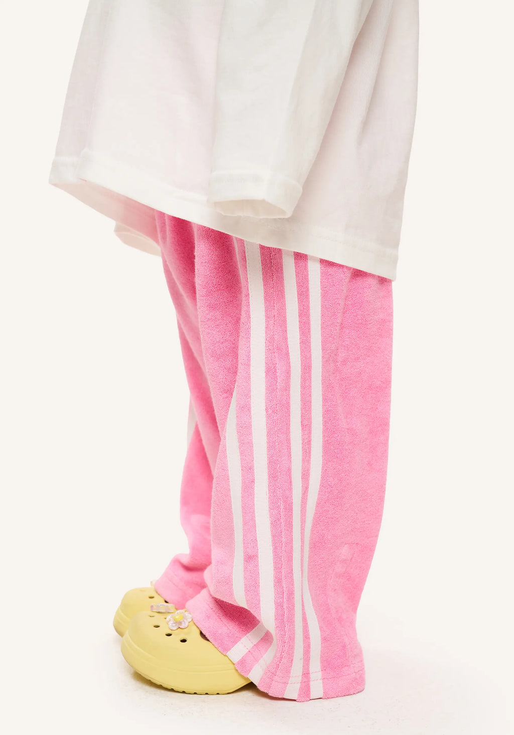 Pink wide-leg sweatpants with three white vertical stripes, styled with a white t-shirt and yellow clogs, shown from the waist down on a light off-white background.