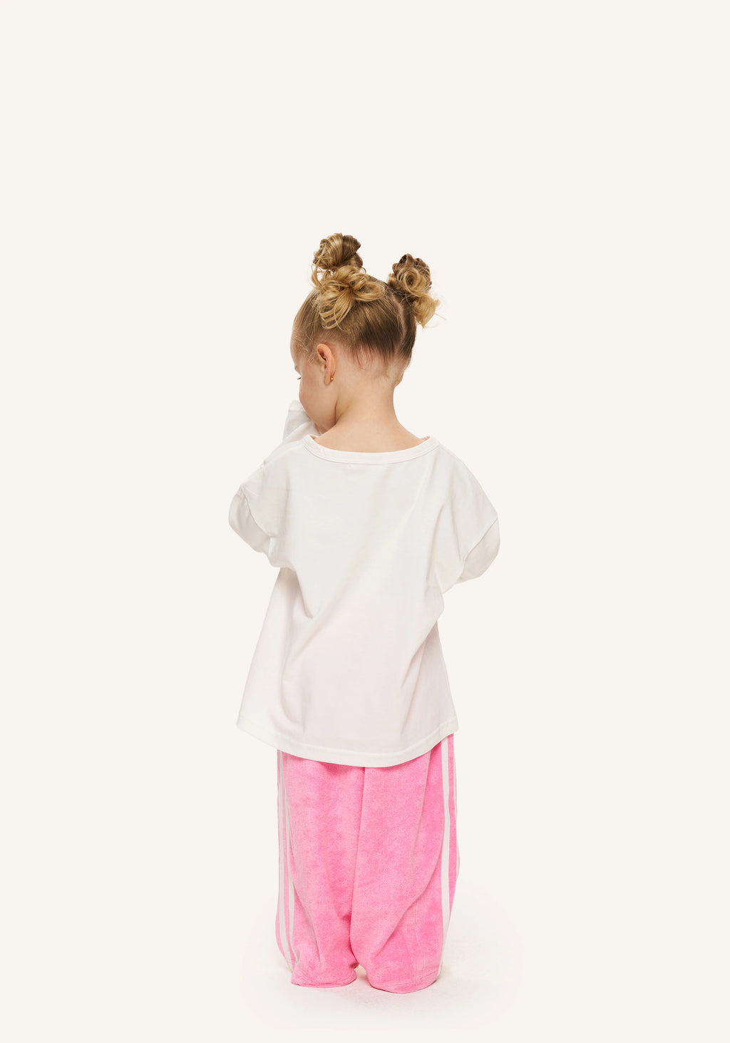 A toddler girl viewed from behind, wearing a white oversized short-sleeve t-shirt and pink wide-leg pants, standing against a solid white background.