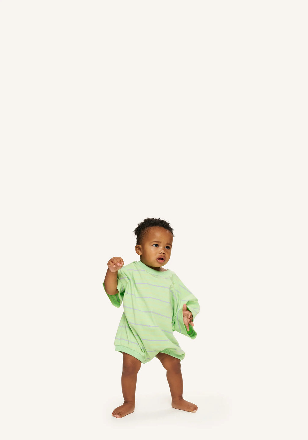A toddler standing barefoot on a plain light background, wearing a light green long-sleeve romper with thin darker green horizontal stripes.