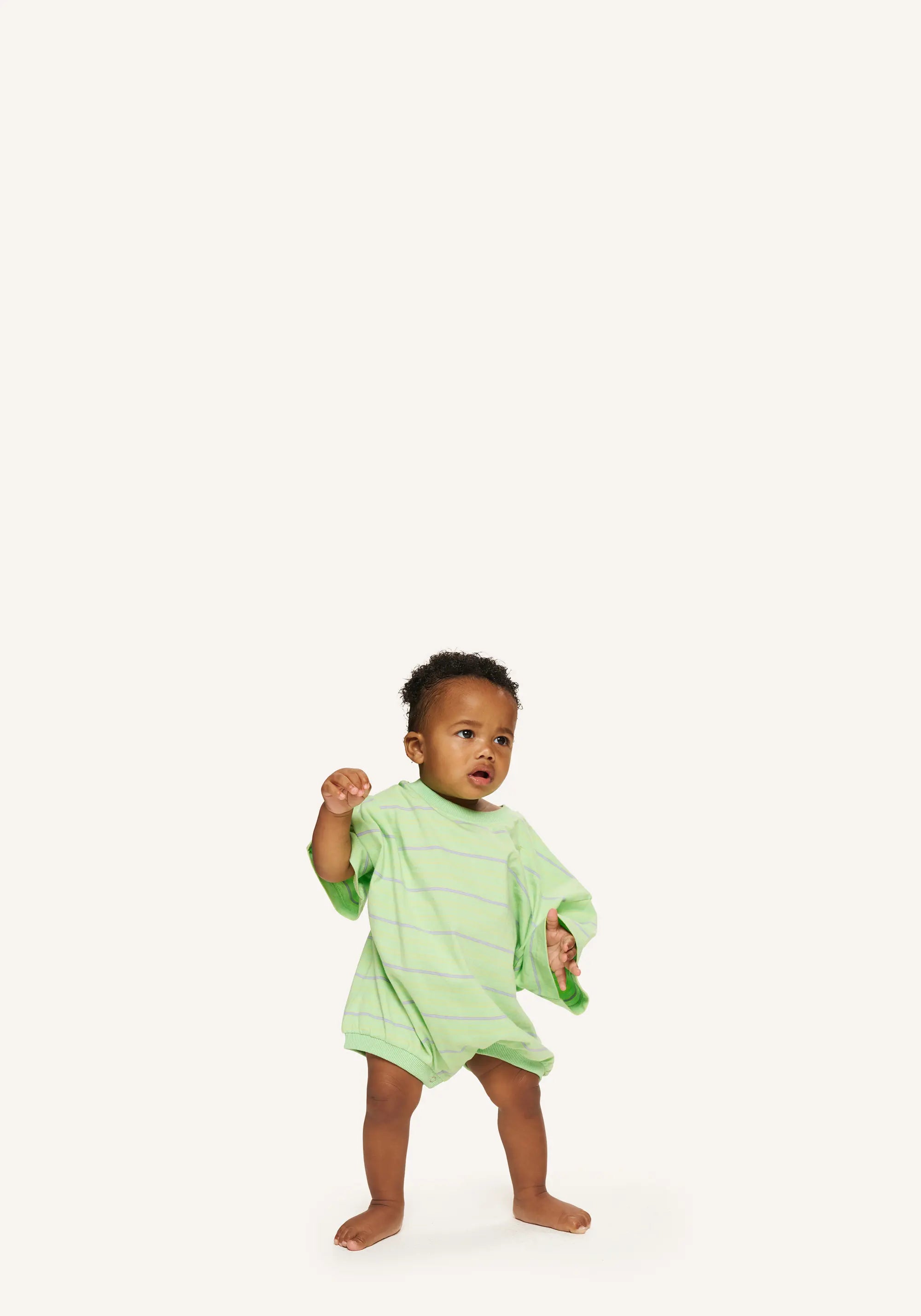 A toddler standing barefoot on a plain light background, wearing a light green long-sleeve romper with thin darker green horizontal stripes.