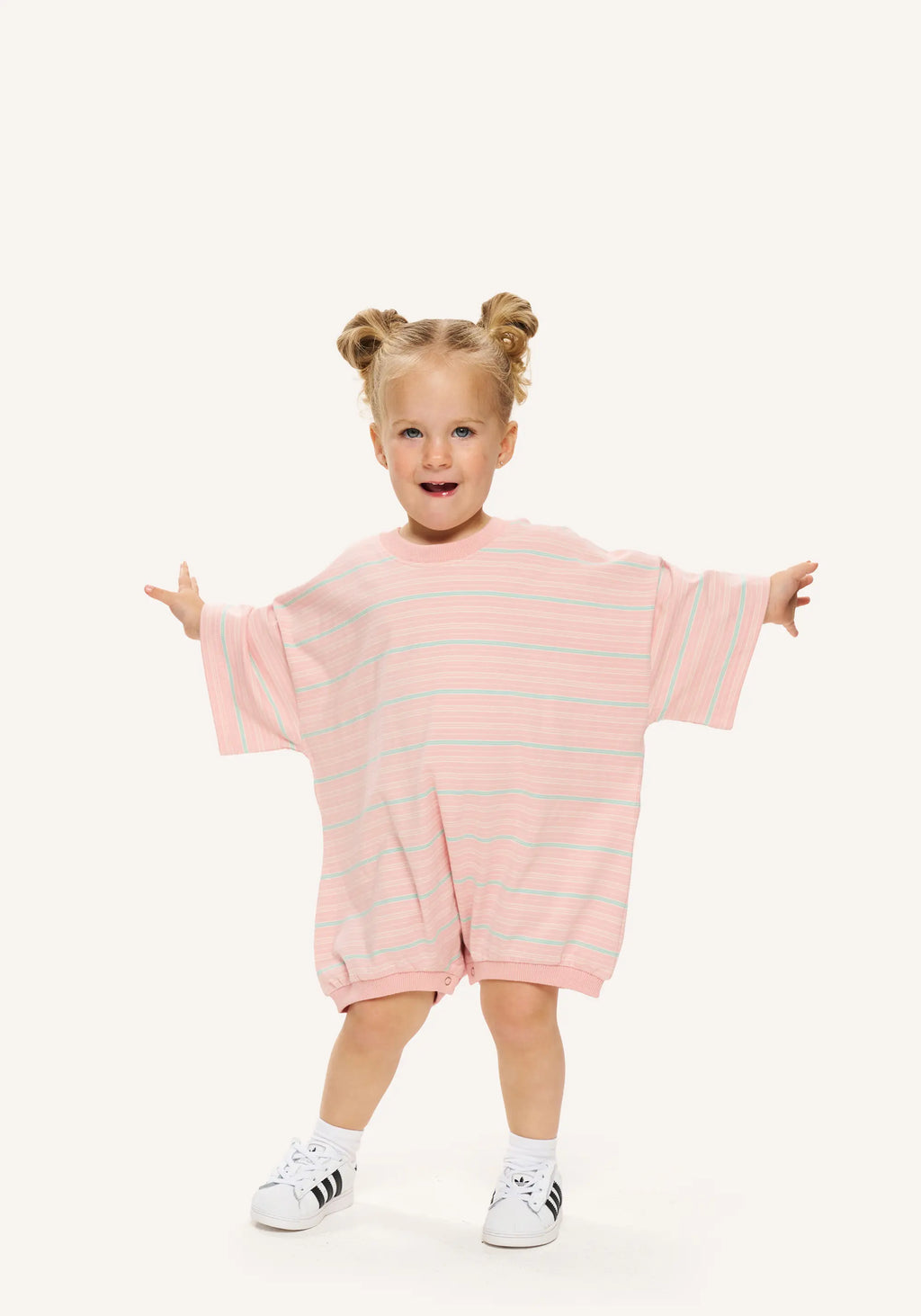 A toddler girl with blonde pigtails stands in a playful pose wearing a pink and light blue striped short-sleeve romper, white socks, and white sneakers against a solid off-white background.