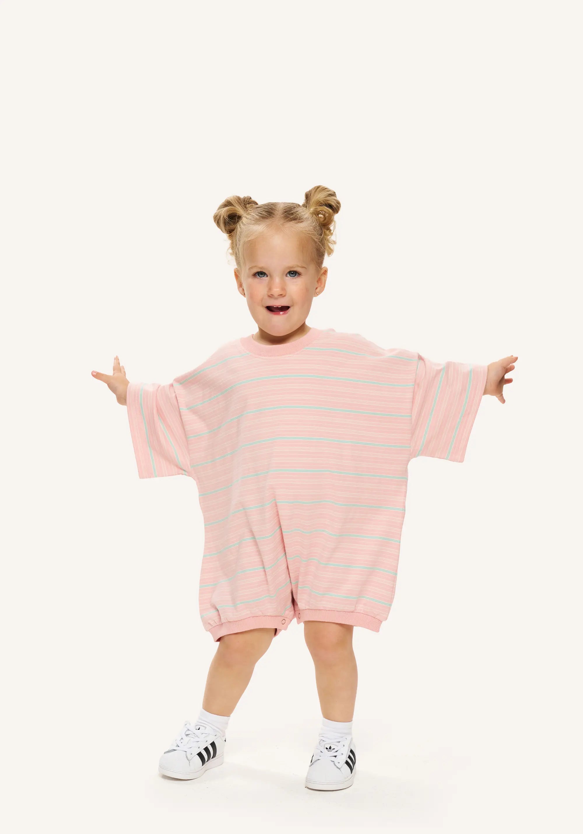 A toddler girl with blonde pigtails stands in a playful pose wearing a pink and light blue striped short-sleeve romper, white socks, and white sneakers against a solid off-white background.