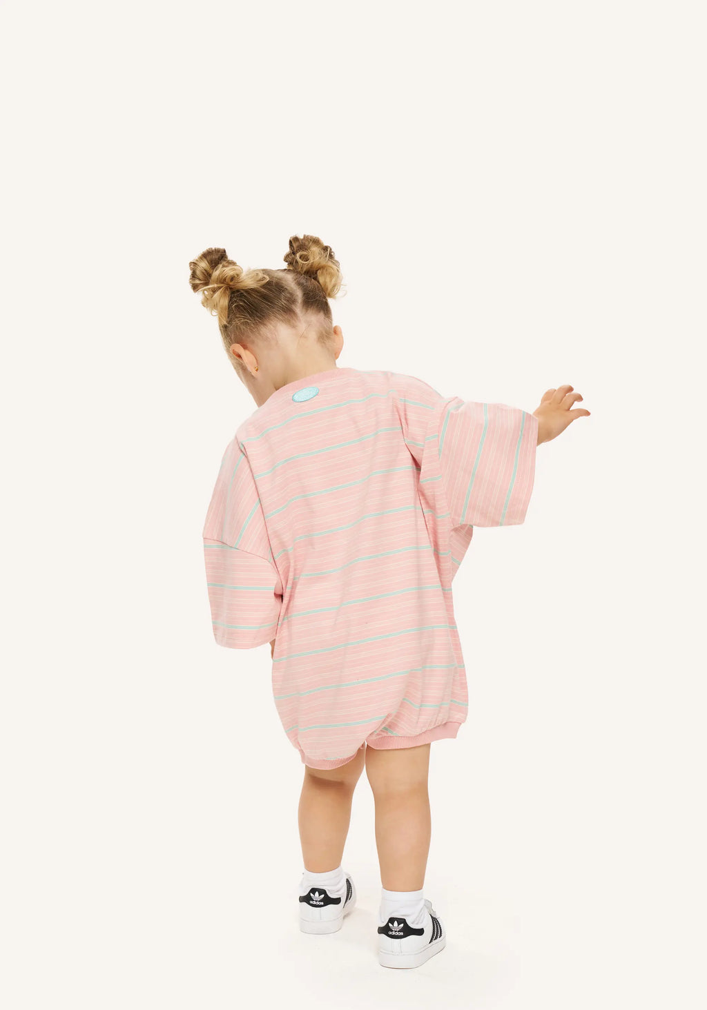 A toddler girl seen from behind wearing a pink and light blue striped long-sleeve romper, standing against a solid off-white background.