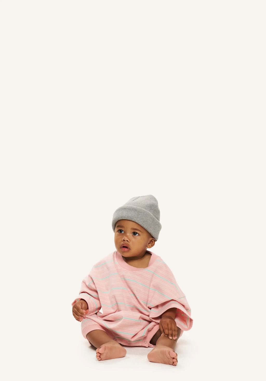 A baby sitting on a white studio background wearing a light pink striped knit romper and a gray beanie hat.