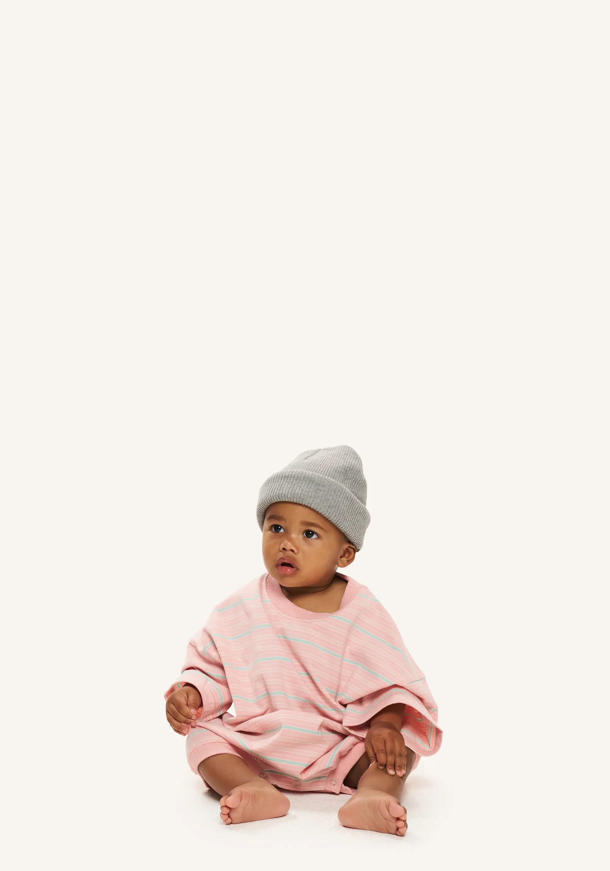 A baby sitting on a white studio background wearing a light pink striped knit romper and a gray beanie hat.