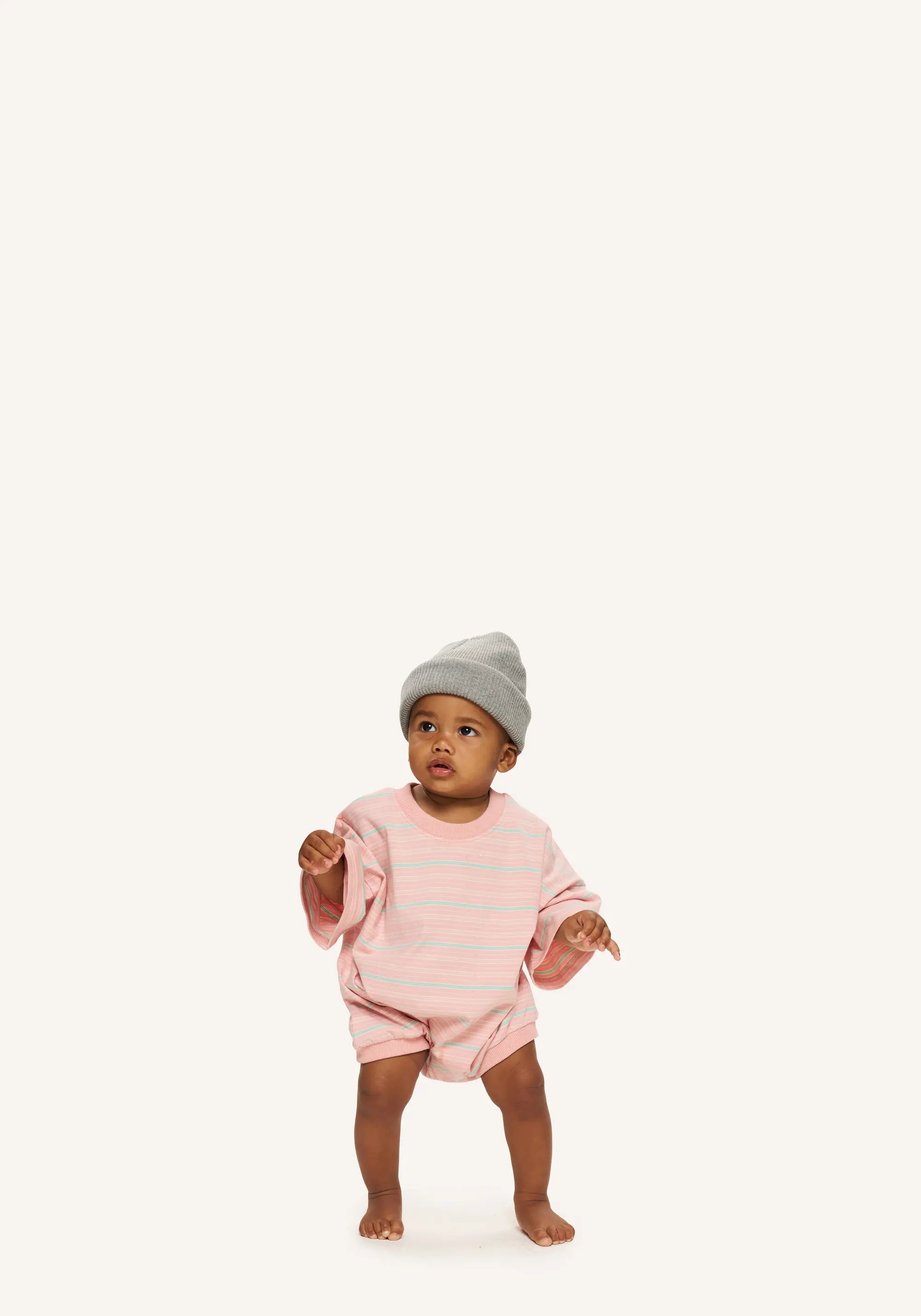 A toddler standing barefoot on a plain off-white background, wearing a light pink and white striped long-sleeve romper and a gray knit beanie.