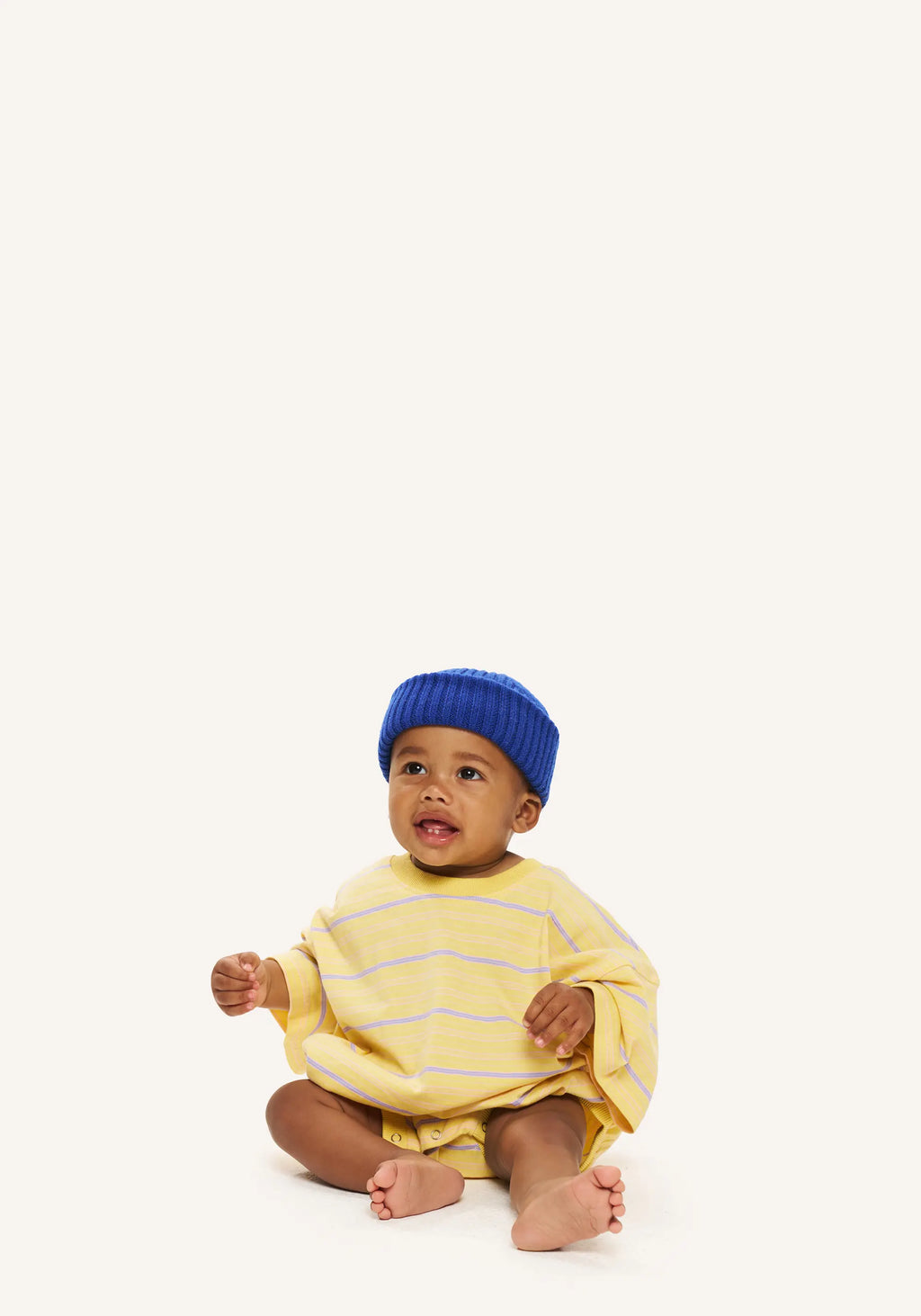 A baby sitting on a white studio floor wearing a yellow and white striped long-sleeve romper and a blue knit beanie hat.