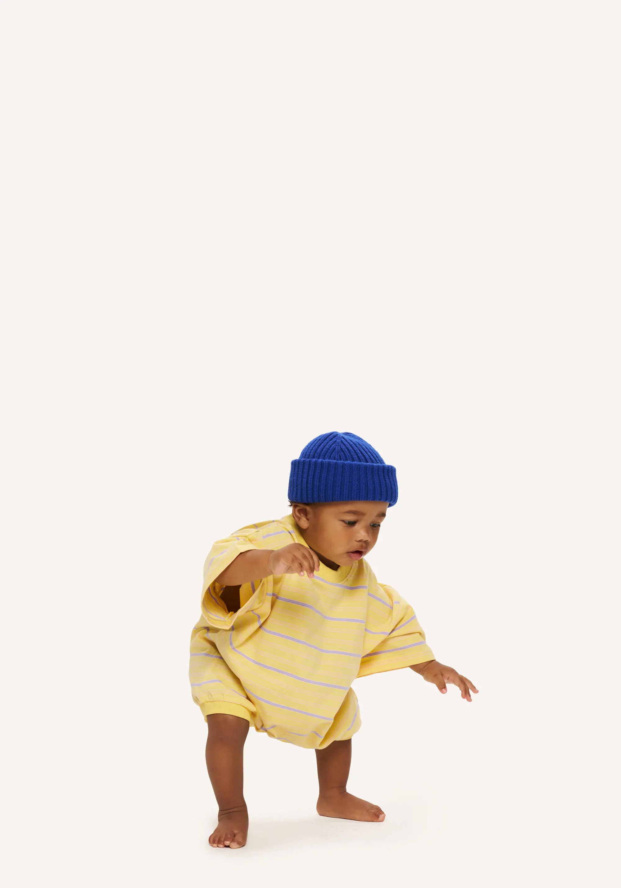 A toddler wearing a yellow and white striped short-sleeve romper and a blue knit beanie hat, captured mid-step against a solid off-white background.