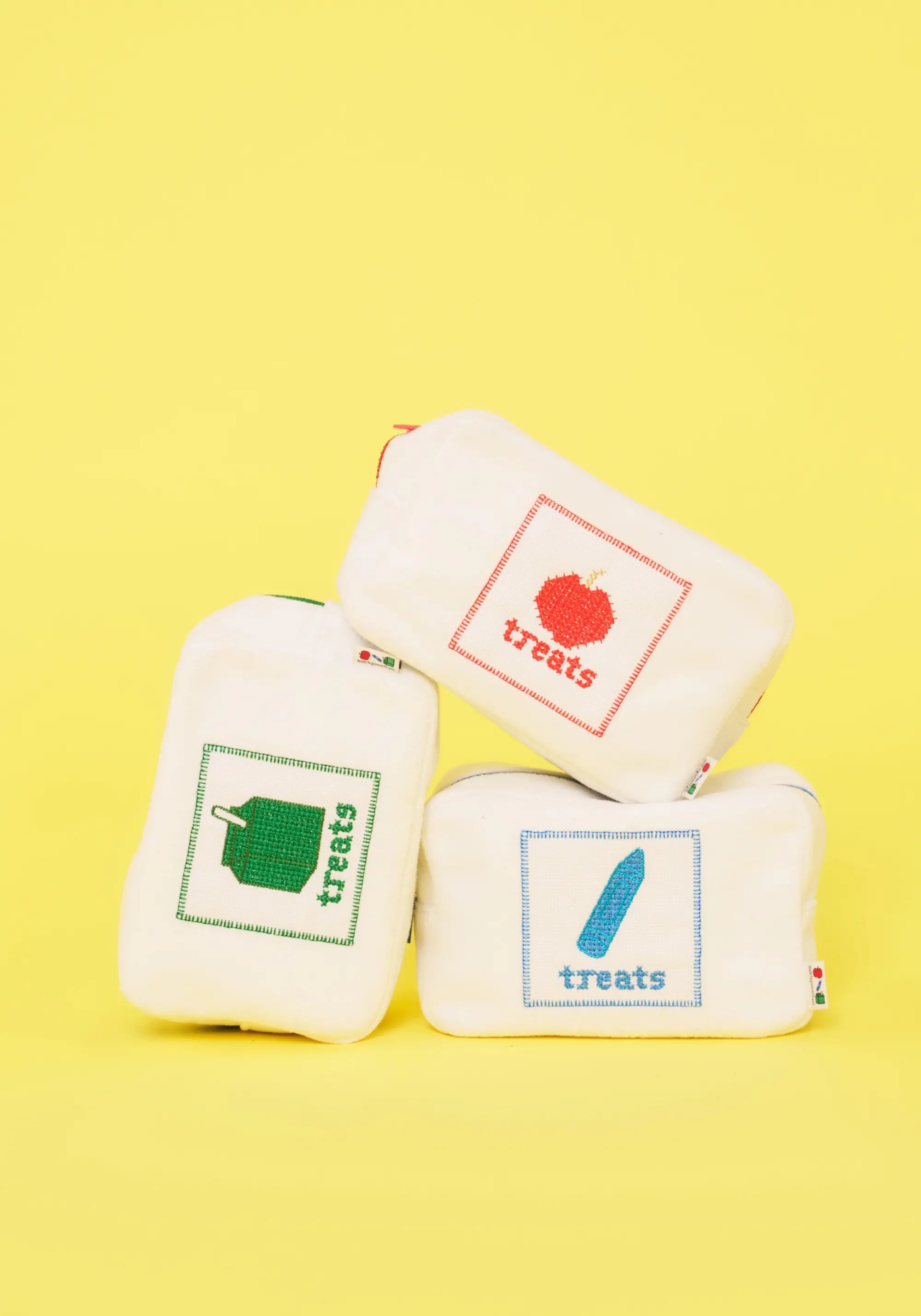 Three small white fabric pouches with colorful embroidered 'treats' graphics, stacked in a pyramid on a bright yellow background.