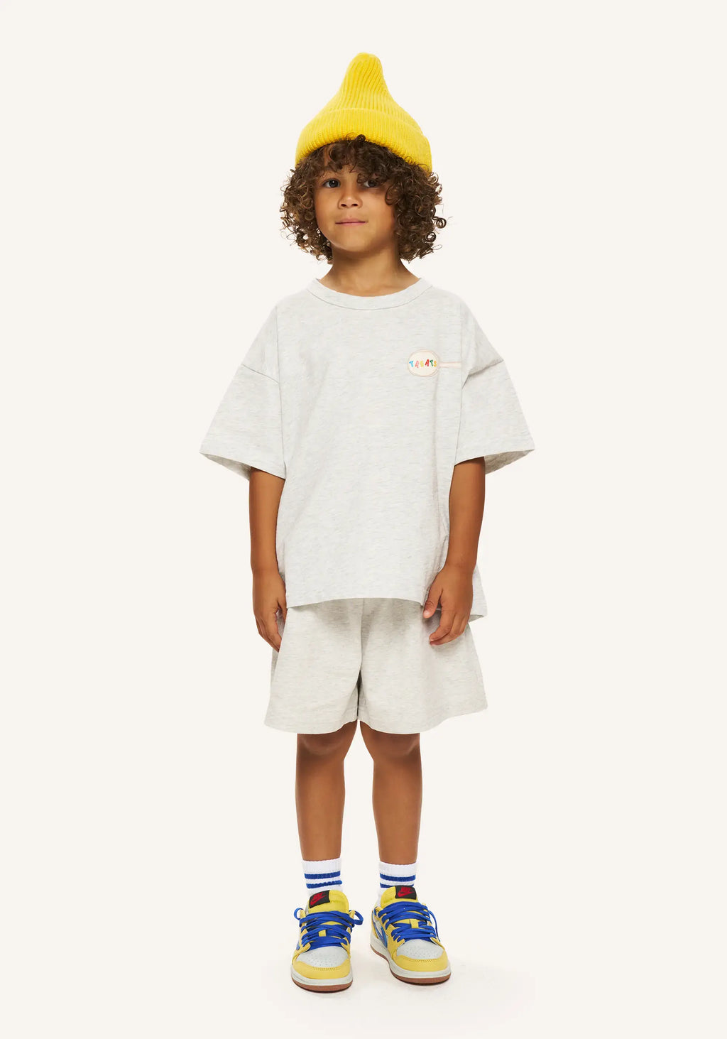 A child model wearing a light gray oversized t-shirt, matching shorts, a bright yellow knit beanie, and colorful sneakers, standing against a solid off-white background.