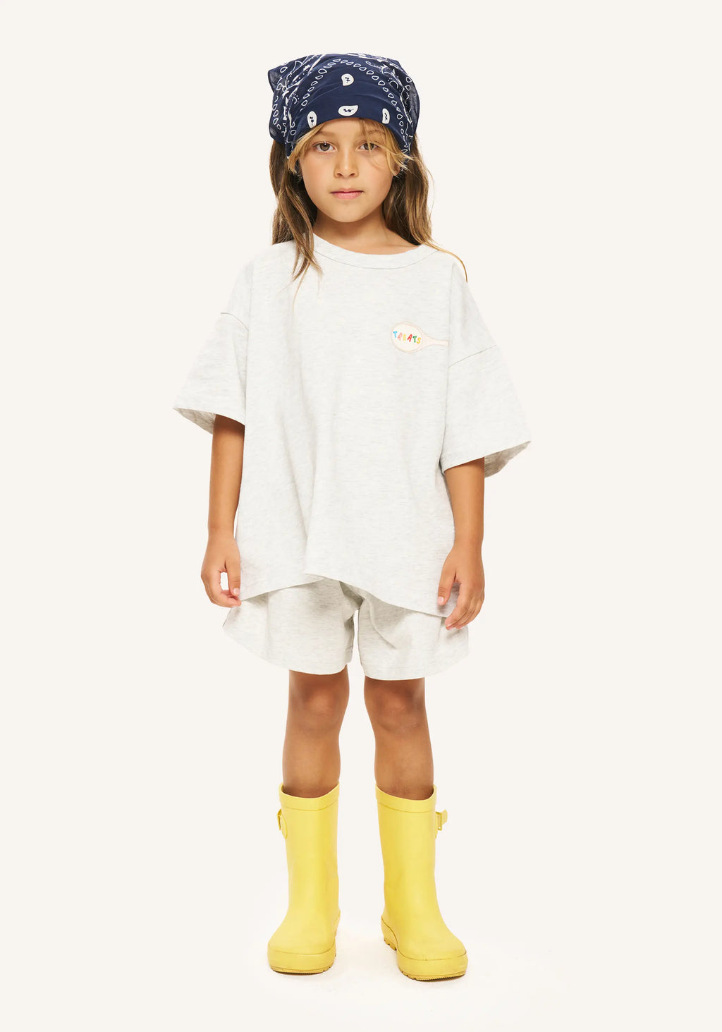 A child model wearing an oversized white t-shirt, matching white shorts, a navy blue patterned bandana, and bright yellow rain boots, standing against a solid off-white background.