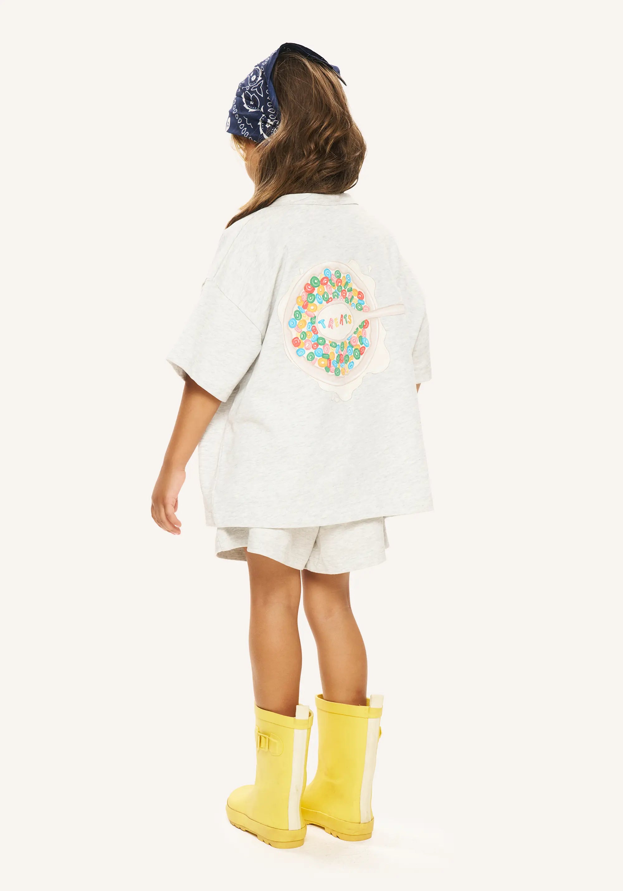 A child shown from the back wearing a light gray oversized T-shirt with a colorful floral graphic on the back, matching shorts, a dark blue patterned headband, and bright yellow rain boots against a plain light background.