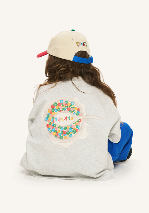 A child seen from behind wearing a light gray sweatshirt with a large colorful cereal graphic on the back, blue pants, and a cream baseball cap with a red brim and blue back strap.