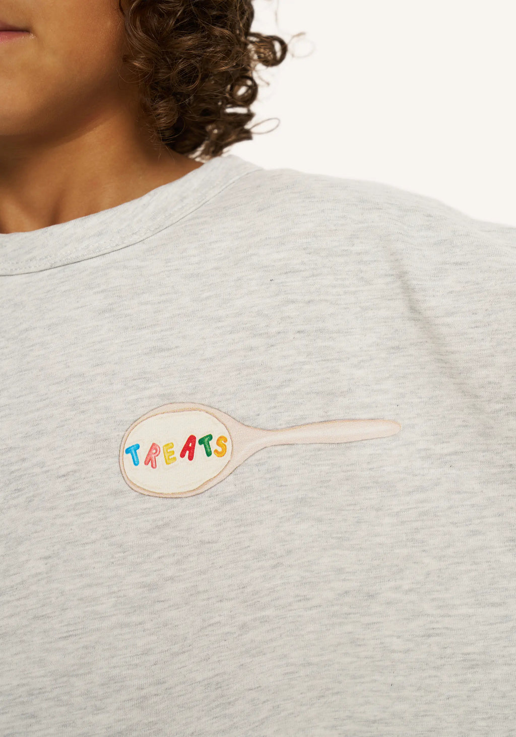 Close-up of a person wearing a light heather gray crewneck sweatshirt. On the chest is a spoon-shaped patch with the word 'TREATS' in colorful letters.