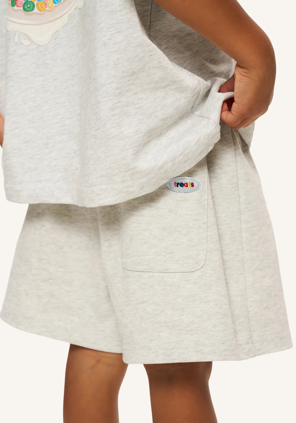 Close-up of a child wearing light heather gray sweatshirt and shorts set, pulling the sweatshirt hem to show a small colorful circular patch with the word 'PROOS' on the side pocket.