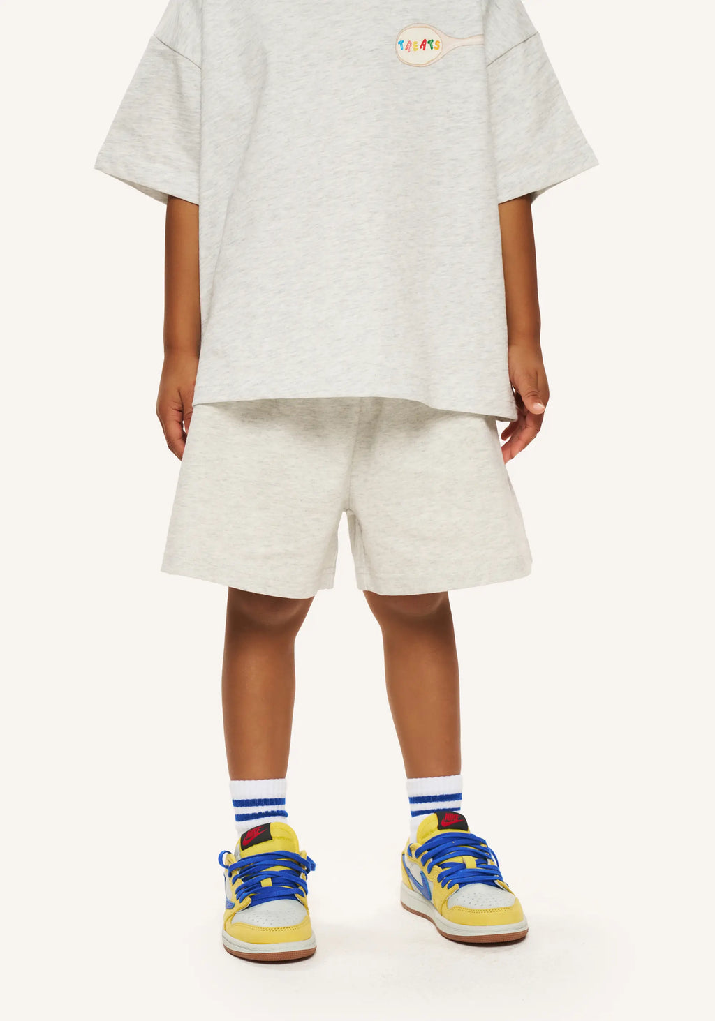 A child model wearing an oversized light gray heather t-shirt and matching shorts, styled with blue and yellow sneakers and striped socks, against a plain off-white background.