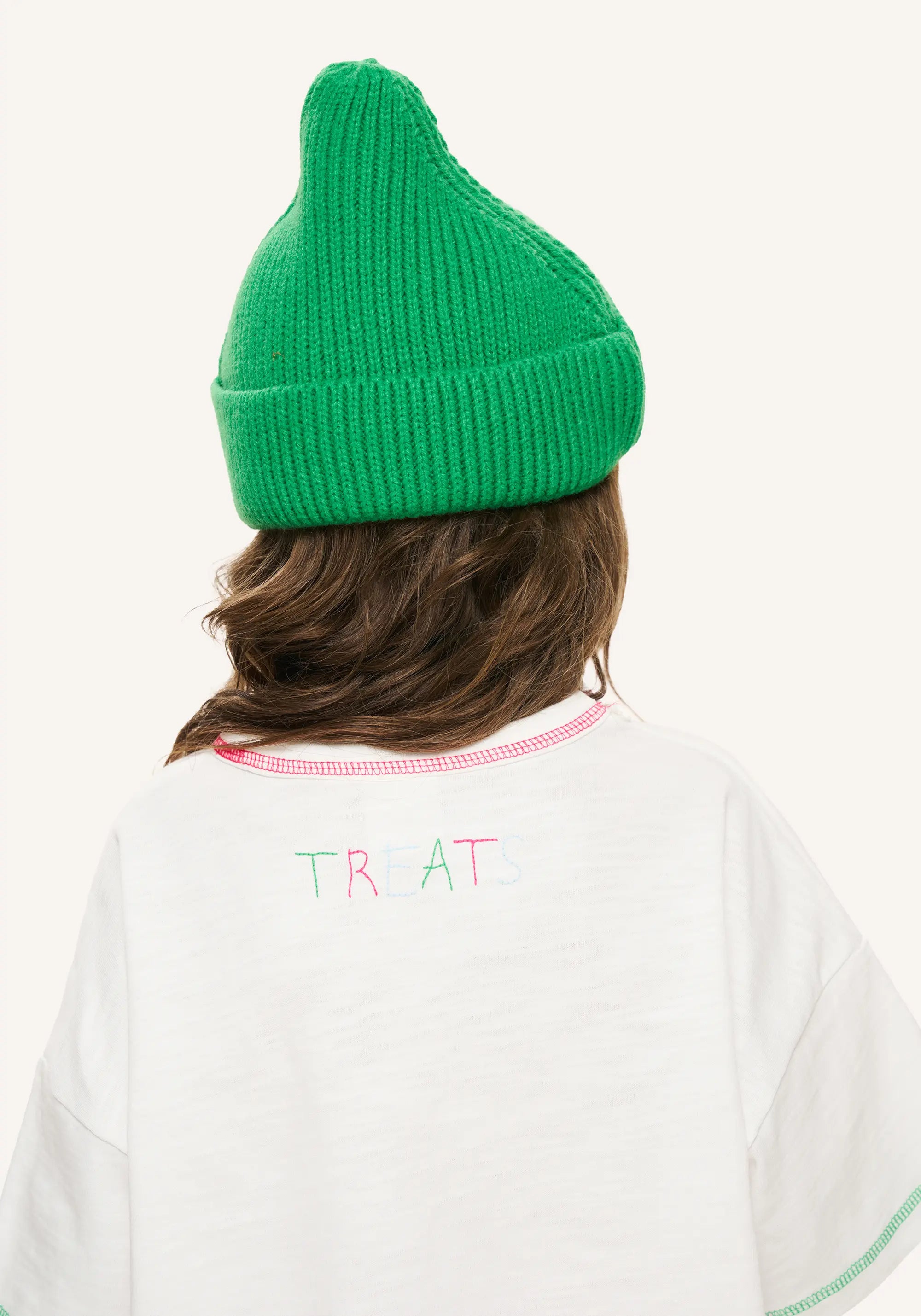 A child shown from behind wearing a bright green ribbed knit beanie and a white T-shirt with 'TREATS' printed in pastel pink and green on the back, against a plain off-white background.