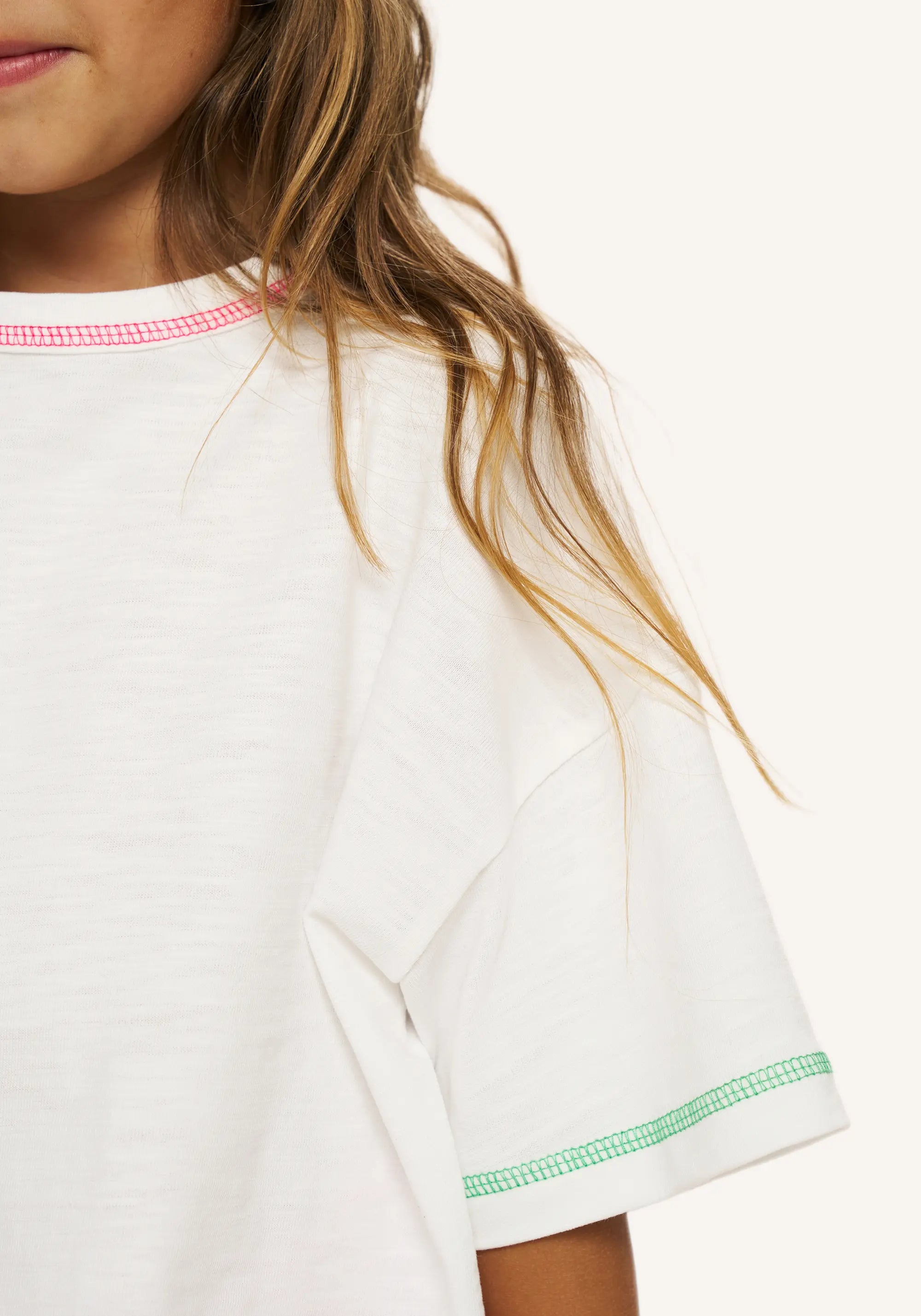 Close-up of a person wearing a white short-sleeve T-shirt with pink gingham trim at the collar and green gingham trim at the sleeve cuff, against a plain light background.