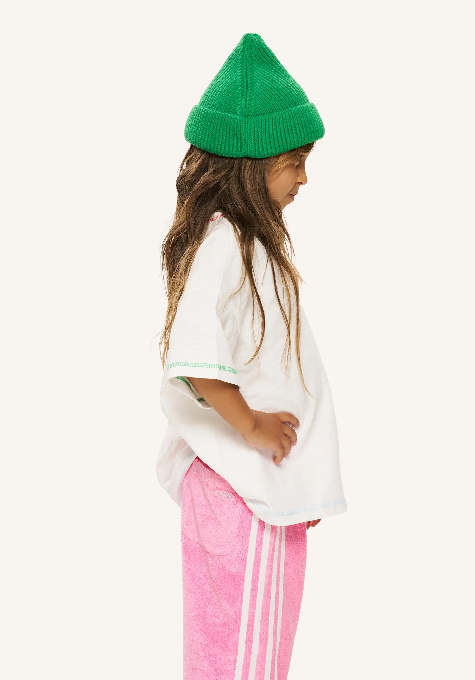 A child model shown in profile wearing a bright green knit beanie, a loose white t-shirt with light green trim, and pink track pants with white side stripes, against a plain off-white background.
