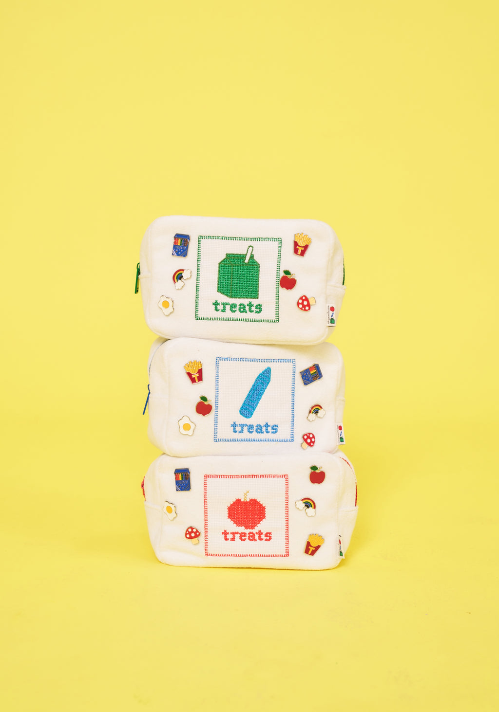 Three small white fabric pouches stacked vertically on a bright yellow background. Each pouch has a different colored square graphic with the word 'treats' and small food illustrations around the edges.