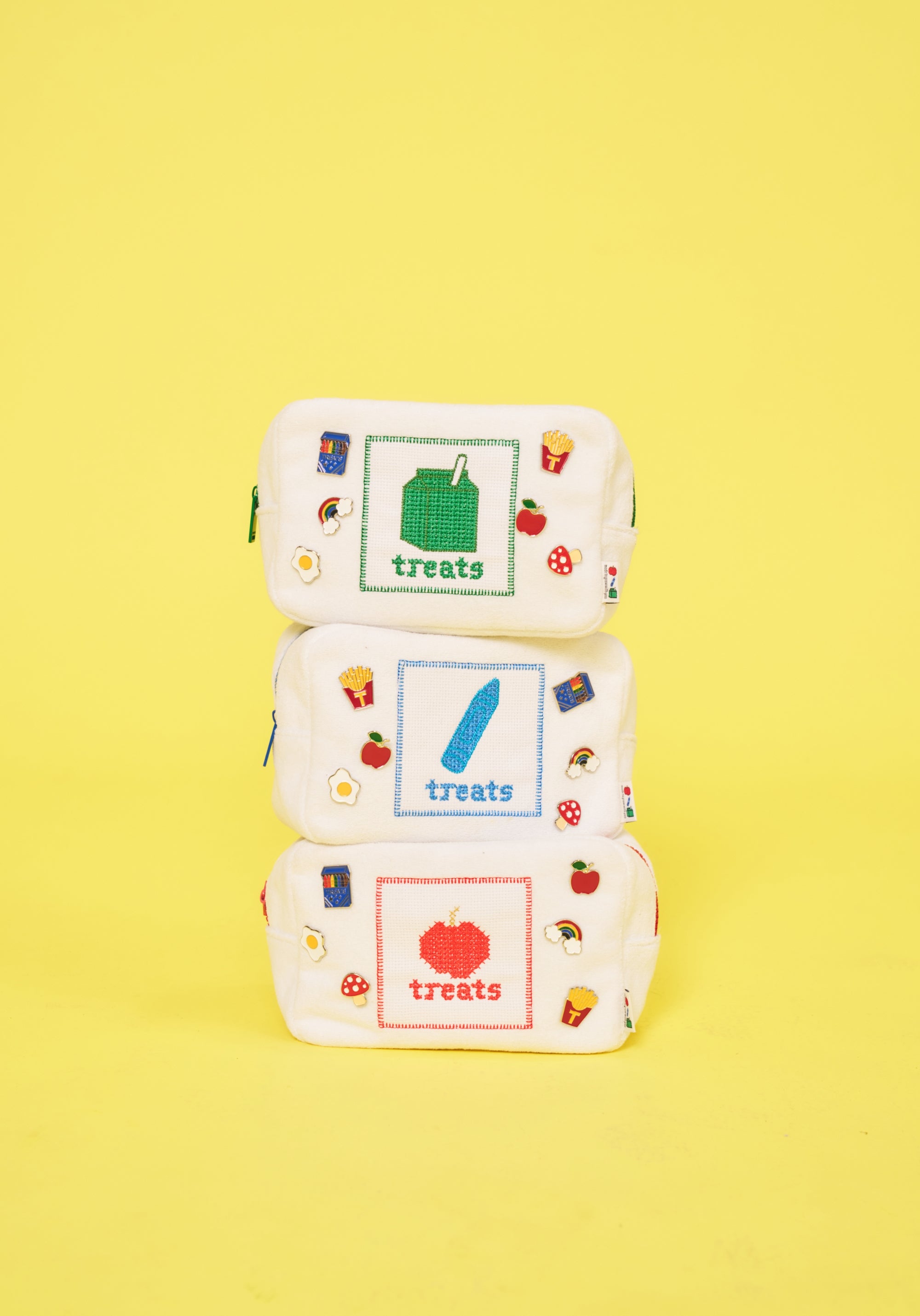Three small white fabric pouches stacked vertically on a bright yellow background. Each pouch has a different colored square graphic with the word 'treats' and small food illustrations around the edges.